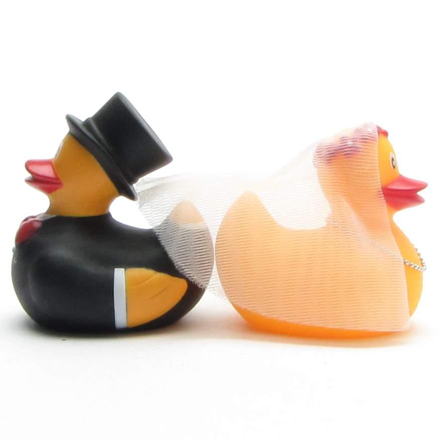 Deux canards en caoutchouc déguisés en mariés, tournés de profil. Le marié porte un smoking noir et un haut-de-forme, tandis que la mariée a un voile blanc sur la tête.