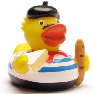Un canard en caoutchouc vêtu d'une tenue française, avec un béret noir, une écharpe rouge et une chemise à rayures bleues, tenant une baguette et un morceau de fromage.