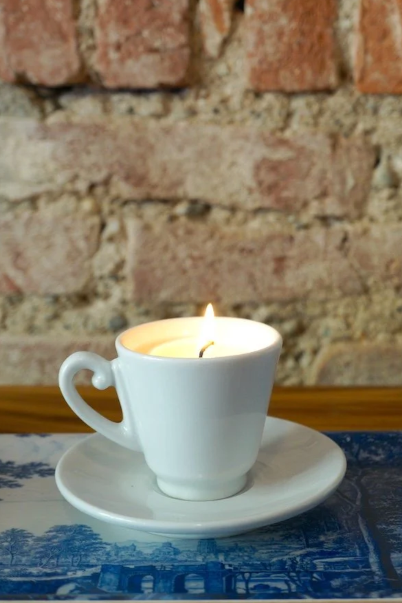Une Candela lumineuse de la collection « tazzina-Festina Lente Milano » est présentée dans une tasse à thé blanche accompagnée de sa soucoupe, disposée sur un plateau à motifs bleus et blancs, avec en arrière-plan un mur de briques au charme rustique.