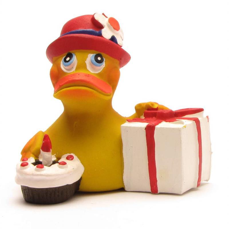 Un canard en caoutchouc jaune, coiffé d'un chapeau rouge orné d'une fleur, tient dans ses mains un coffret cadeau blanc orné d'un ruban rouge, ainsi qu'un petit gâteau surmonté d'une bougie.