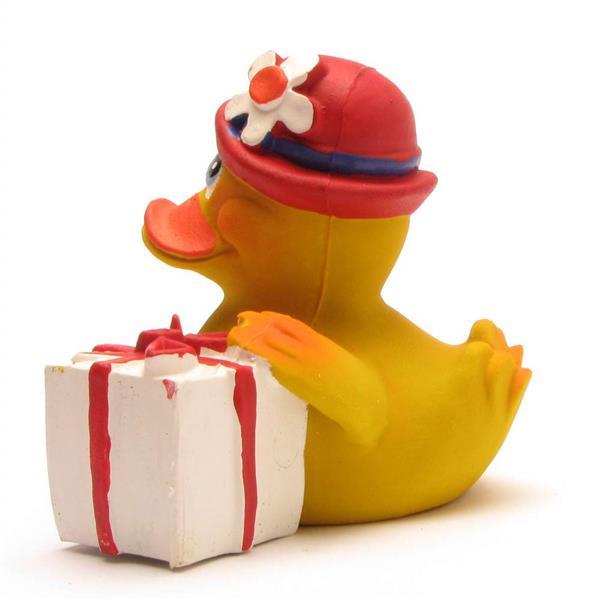 Un canard en caoutchouc jaune coiffé d'un chapeau rouge orné d'une fleur blanche, assis à côté d'un coffret cadeau blanc noué d'un ruban rouge.