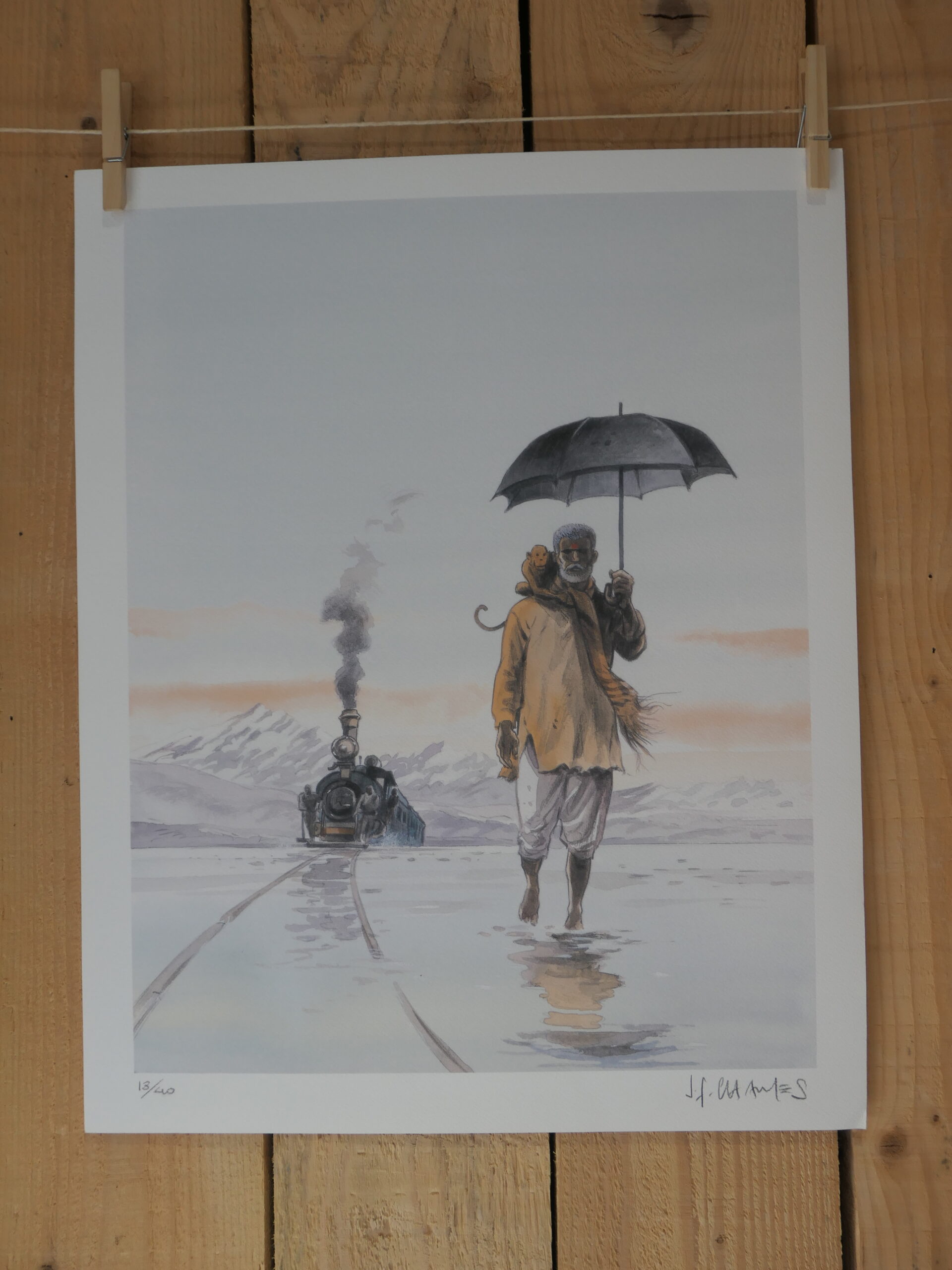 Une lithographie intitulée « L’Homme au parapluie » de Jean-François Charles (40 × 50 cm) représente un personnage tenant un parapluie qui marche sur des rails mouillés en direction du spectateur, avec un train à vapeur qui approche derrière lui. Des montagnes enneigées et un ciel nuageux constituent l’arrière-plan, et l’œuvre est accrochée à un mur en bois.