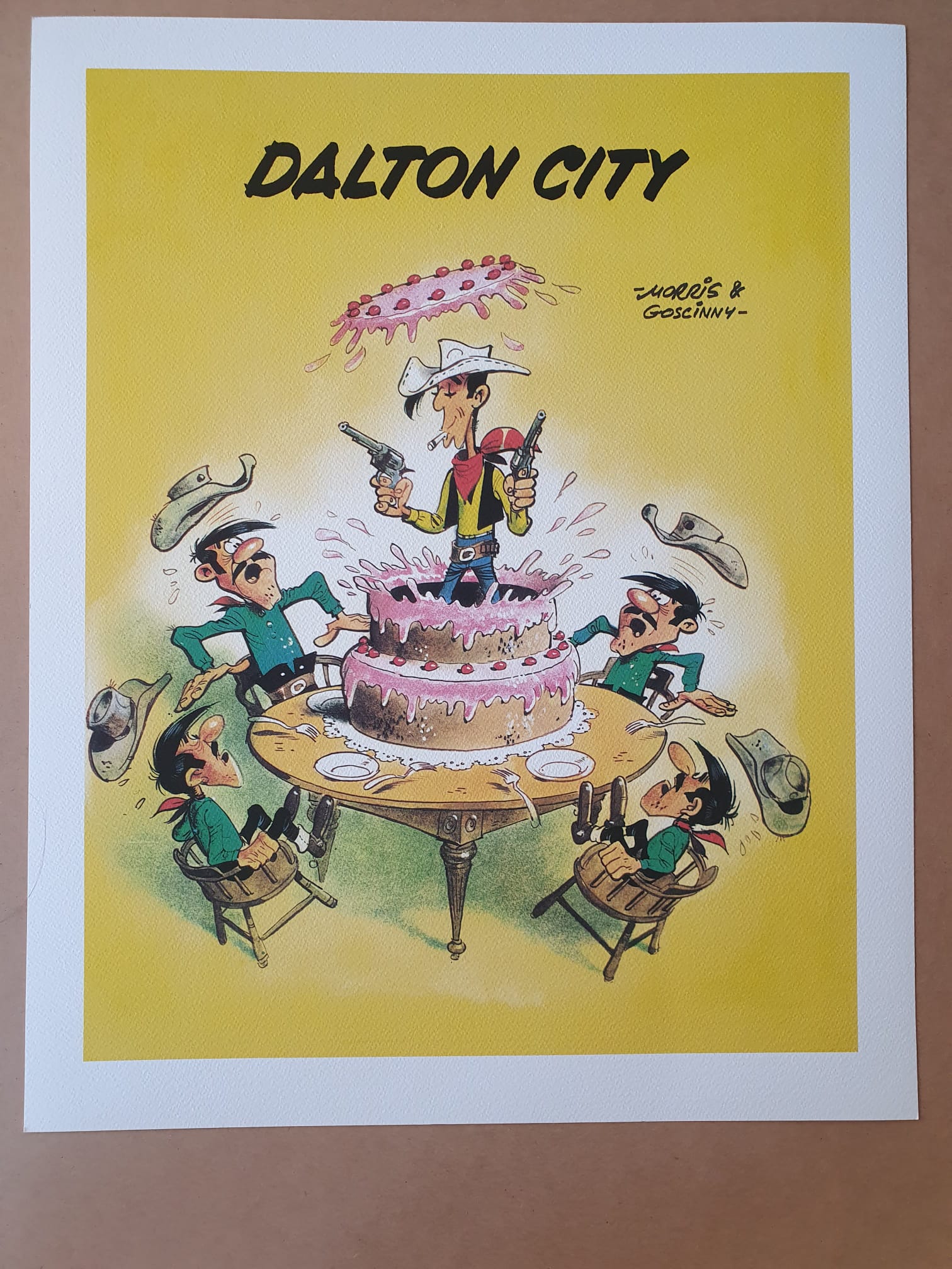 Une lithographie intitulée « Lithographie Morris – Lucky Luke : Dalton City – 40 × 50 cm », représentant une illustration humoristique d’un cow-boy armé de deux pistolets, debout au milieu d’un gâteau écrasé posé sur une table, entouré de quatre hommes surpris tombant de leurs chaises, avec l’inscription « DALTON CITY » au-dessus.