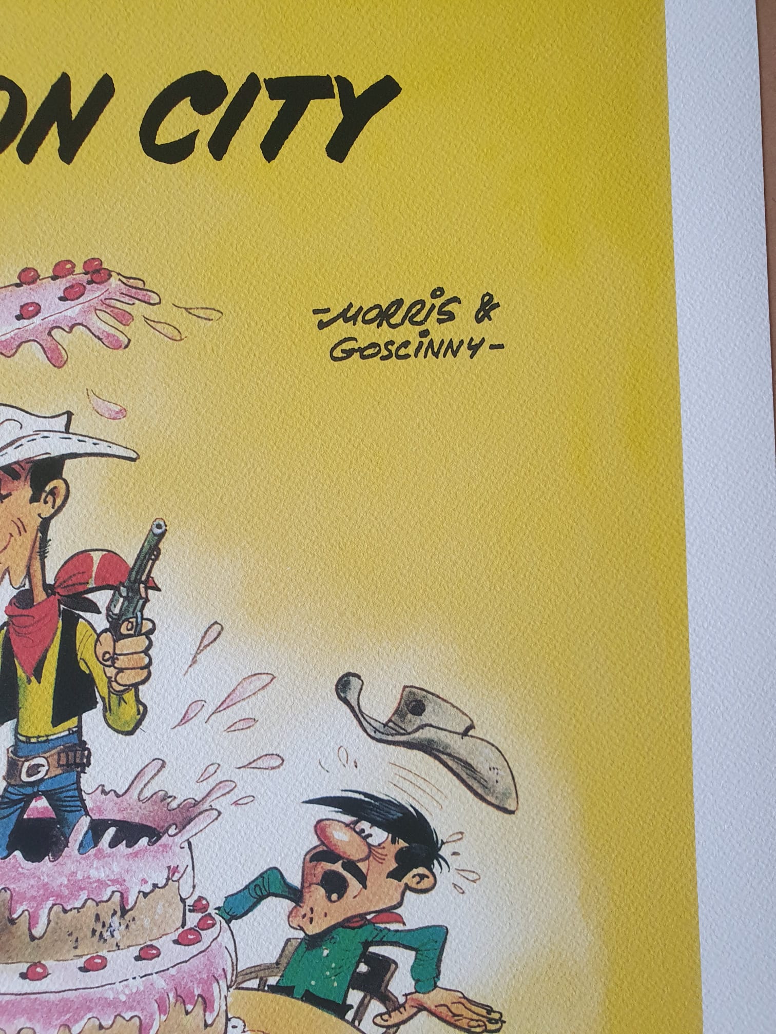 La lithographie de Morris – Lucky Luke : Dalton City – 40 × 50 cm représente un cow-boy de bande dessinée tenant un pistolet et debout au milieu d'un gâteau rose, surprenant un homme moustachu qui tombe à la renverse et perd son chapeau, avec la mention « Morris & Goscinny » inscrite sur le fond jaune.