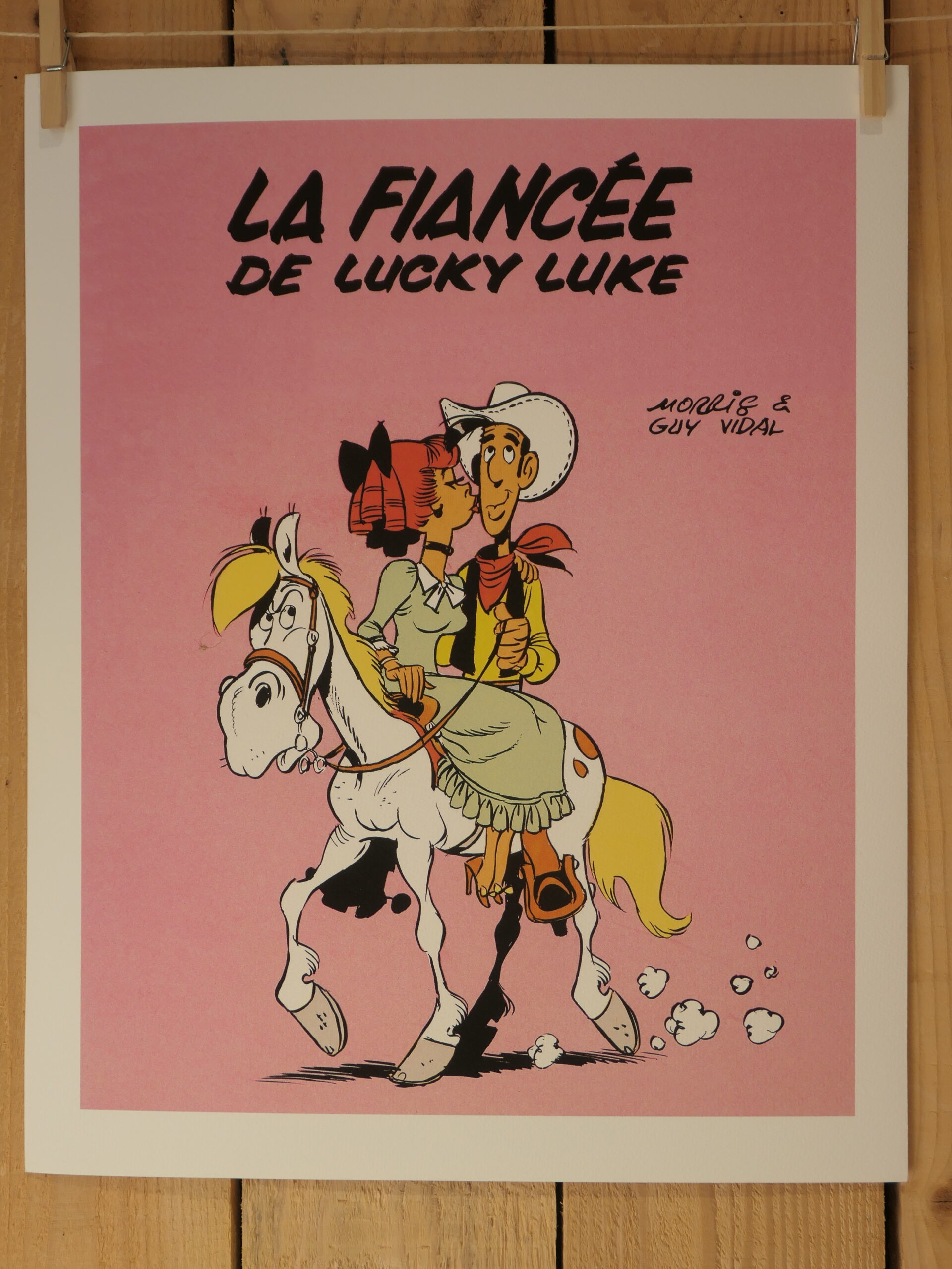 Une lithographie intitulée « Lithographie Morris – Lucky Luke : La Fiancée – 40 × 50 cm » représente Lucky Luke à cheval aux côtés d’une femme vêtue d’une robe verte, avec le titre « La Fiancée de Lucky Luke » apparaissant au-dessus d’eux sur un fond rose ; les noms des auteurs, Morris et Guy Vidal, figurent à droite.