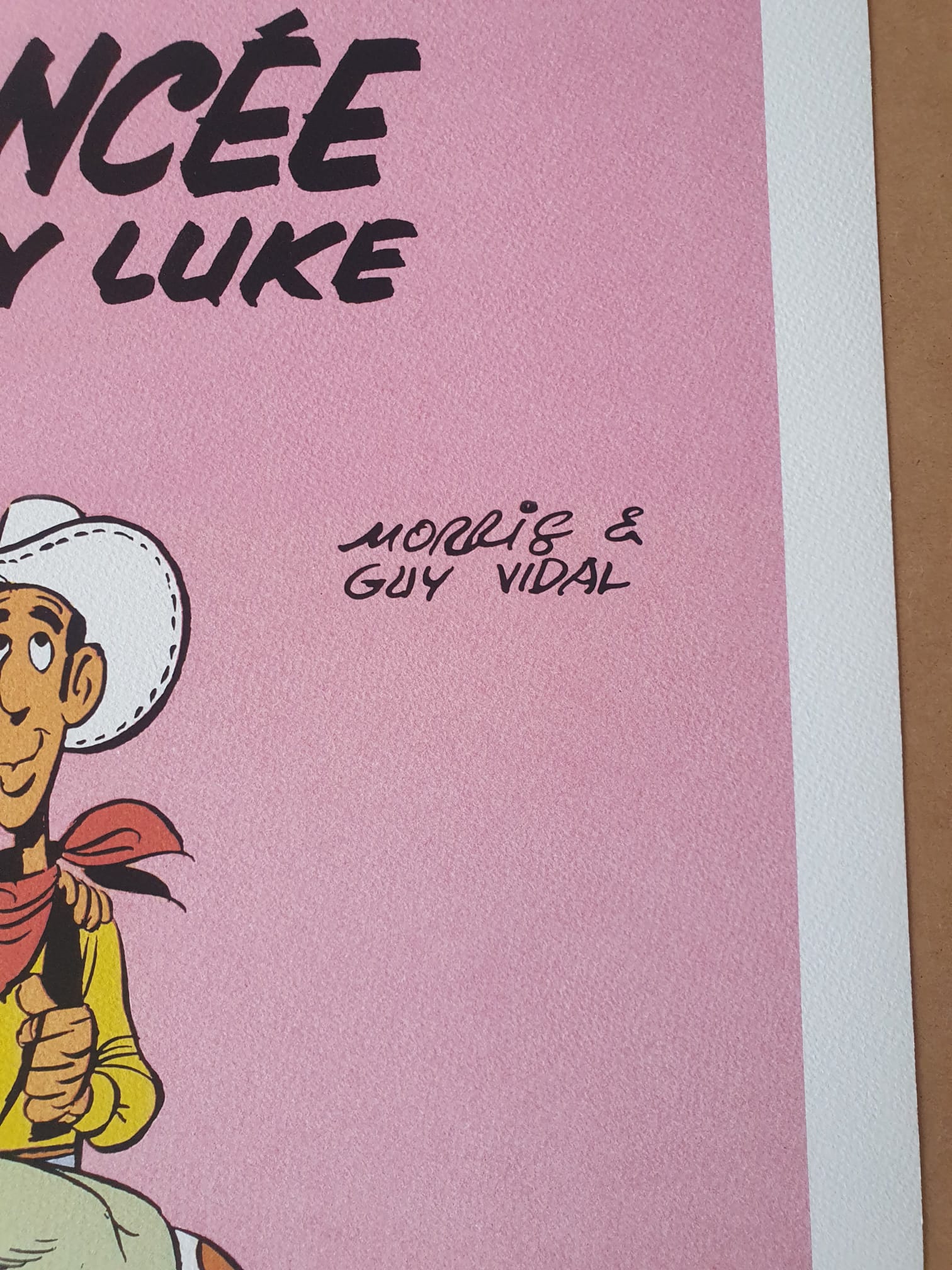 Un gros plan de l'illustration de bande dessinée « Lithographie Morris – Lucky Luke : La Fiancée – 40 × 50 cm » montre une partie du personnage de cow-boy Lucky Luke sur un fond rose, avec la mention manuscrite « Morris & Guy Vidal » près du centre.