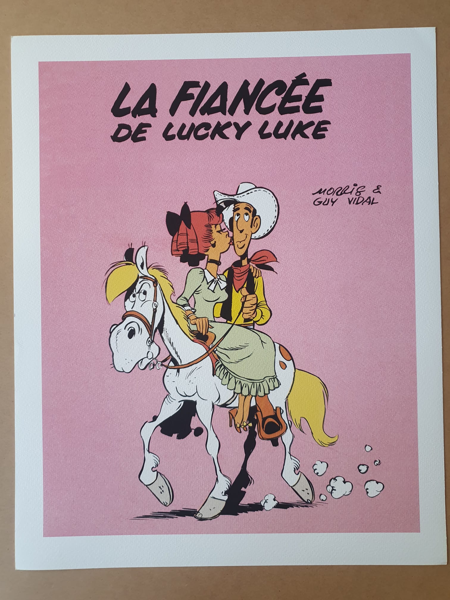 Lithographie Morris – Lucky Luke : La Fiancée – 40 × 50 cm représente Lucky Luke en tenue de cow-boy, chevauchant un cheval blanc, avec une femme en robe rose assise derrière lui. L'arrière-plan est rose et comporte les inscriptions « La Fiancée de Lucky Luke » et « Morris & Guy Vidal ».