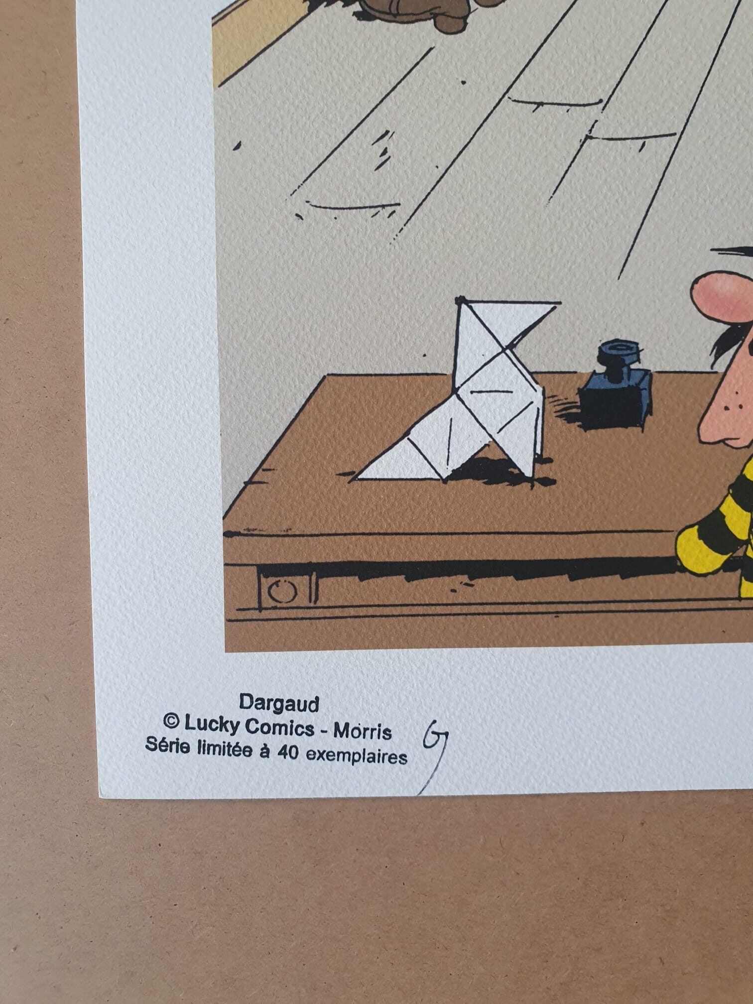 Un gros plan de la lithographie « Morris – Lucky Luke : Marcel Dalton – 40 × 50 cm » montre un coin de l'estampe sur lequel figurent une signature, le logo Dargaud, les mentions de droits d'auteur de « Lucky Comics - Morris », ainsi qu'une mention indiquant que l'édition est limitée à 40 exemplaires. On aperçoit également une partie d'une illustration de bande dessinée et un bureau en bois.