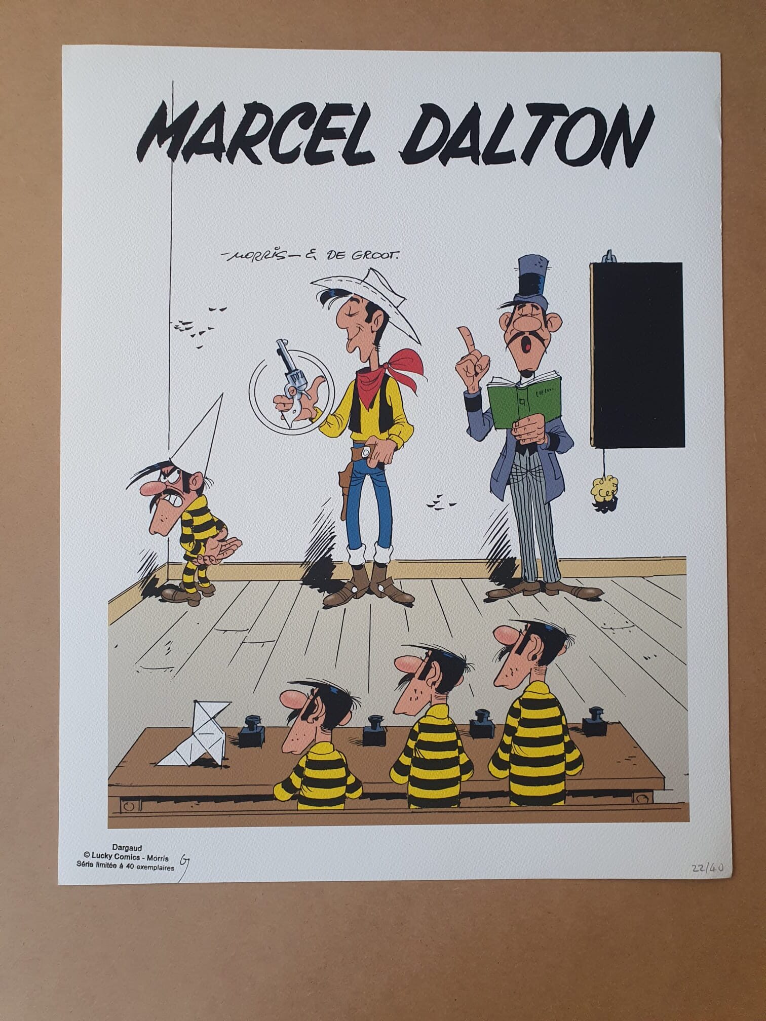 Illustration de style bande dessinée intitulée « Lithographie Morris – Lucky Luke : Marcel Dalton – 40 × 50 cm », représentant quatre hommes en tenues rayées jaunes et noires assis à des bureaux, un homme en blanc qui pointe du doigt, et un homme de grande taille coiffé d'un haut-de-forme tenant un livre devant un tableau noir. Le nom « Marcel Dalton » apparaît en haut de l'illustration.