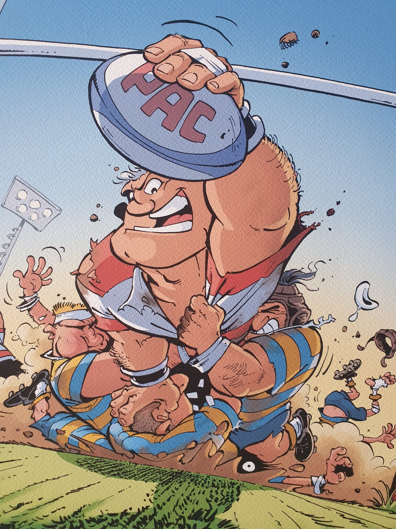 Un joueur de rugby de bande dessinée, un large sourire aux lèvres, tient au-dessus de sa tête un ballon portant l’inscription « PAC » tout en esquivant des adversaires vêtus d’uniformes bleus et jaunes, tandis que la terre et l’herbe volent à mesure qu’ils se bousculent — illustration tirée de la lithographie de Jean-Charles Poupard intitulée « Rugby 2 » (40 × 50 cm).