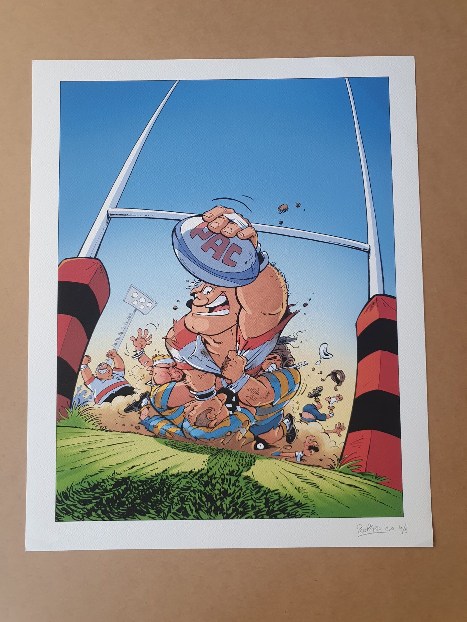 Une scène animée tirée de la « Lithographie Jean-Charles Poupard – Rugby 2 – 40 × 50 cm » représente un joueur de rugby caricatural, vêtu de bleu et de jaune, marquant un essai, le ballon marqué « FAC » serré dans les mains, tandis que ses coéquipiers et ses adversaires se bousculent en contrebas, avec les poteaux de but et des supporters en liesse en arrière-plan.