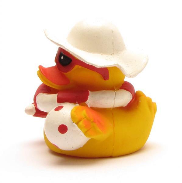 Un canard en caoutchouc jaune porte un chapeau de soleil blanc et des lunettes de soleil rouges ; il tient un ballon de plage blanc à pois rouges et est assis sur un fond blanc.