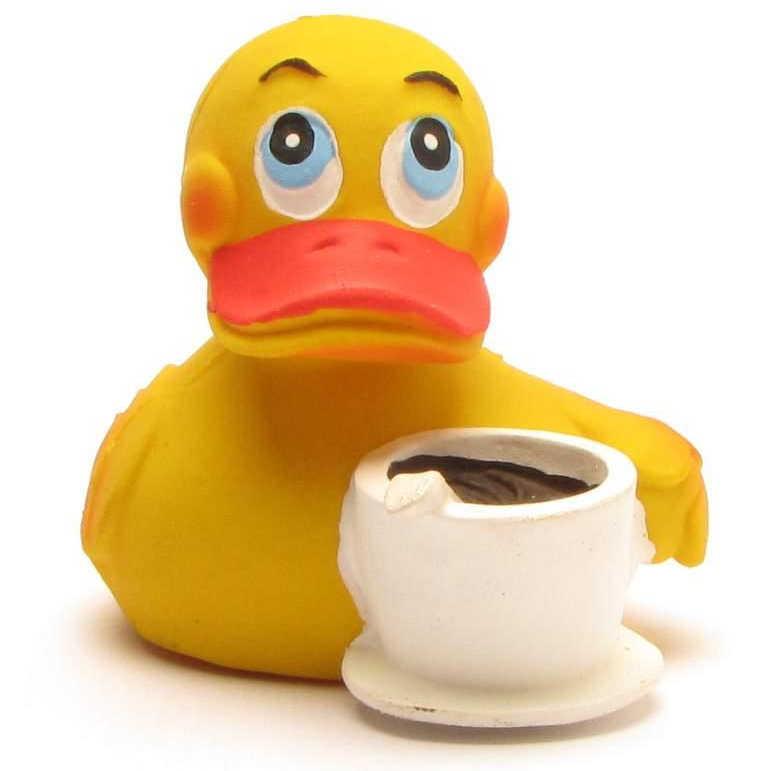 Un canard en caoutchouc jaune aux yeux bleus est posé à côté d'une tasse à café blanche et de sa soucoupe ; on dirait qu'il tient la tasse avec une aile.