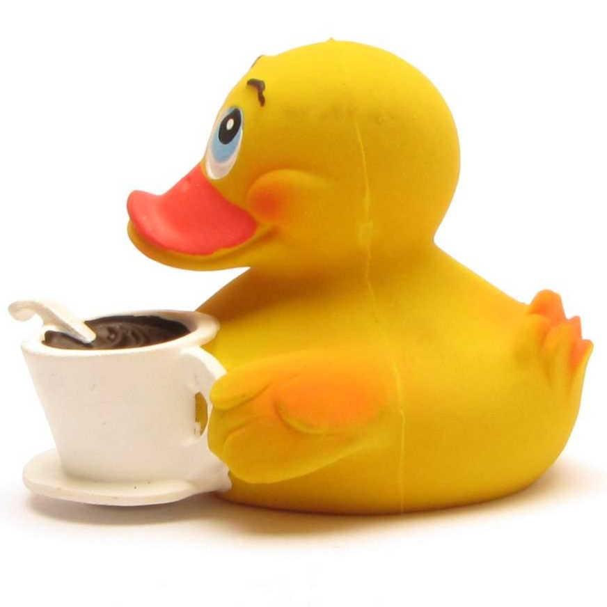 Un canard en caoutchouc jaune tient une tasse de café blanche dans laquelle se trouve une cuillère ; il a l'air joyeux et satisfait.