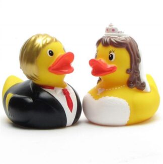 Deux canards en caoutchouc déguisés en mariés se font face. Le canard marié porte un costume et une cravate rouge, tandis que la canarde mariée porte une robe blanche, un collier, un diadème et a les cheveux bruns.