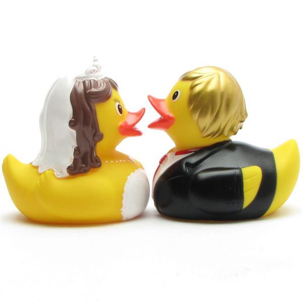 Deux canards en caoutchouc déguisés en mariés se font face. La mariée porte un voile et une robe blancs, tandis que le marié porte un smoking noir avec un nœud papillon rouge et a les cheveux jaunes.