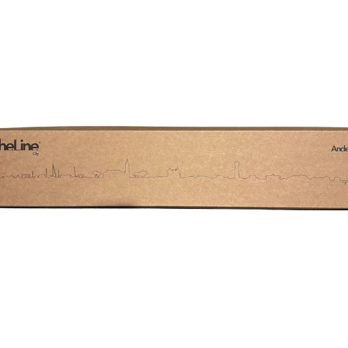 Une boîte en carton marron est ornée, sur toute sa longueur, d’un dessin au trait noir simple et fin représentant la silhouette de la ville d’Andernos-les-Bains. La boîte comporte également une partie du logo « Skyline murale Andernos-les-Bains en métal noir – 30 cm » ainsi que du texte dans les coins supérieurs gauche et droit.