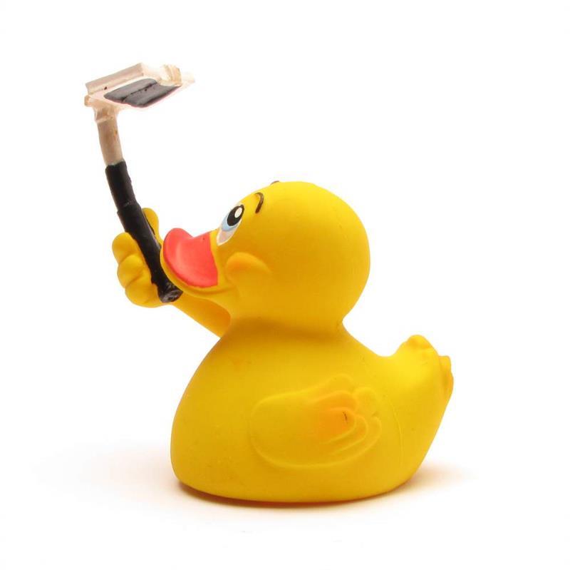 Un canard en caoutchouc jaune tient une perche à selfie noire à laquelle est fixé un smartphone, comme s'il prenait un selfie, sur un fond blanc.