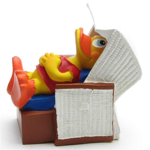 Un canard de dessin animé jaune, vêtu d'une tenue rouge, est allongé sur le dos sur un coussin bleu, se détendant dans une chaise longue en osier marron et blanche.