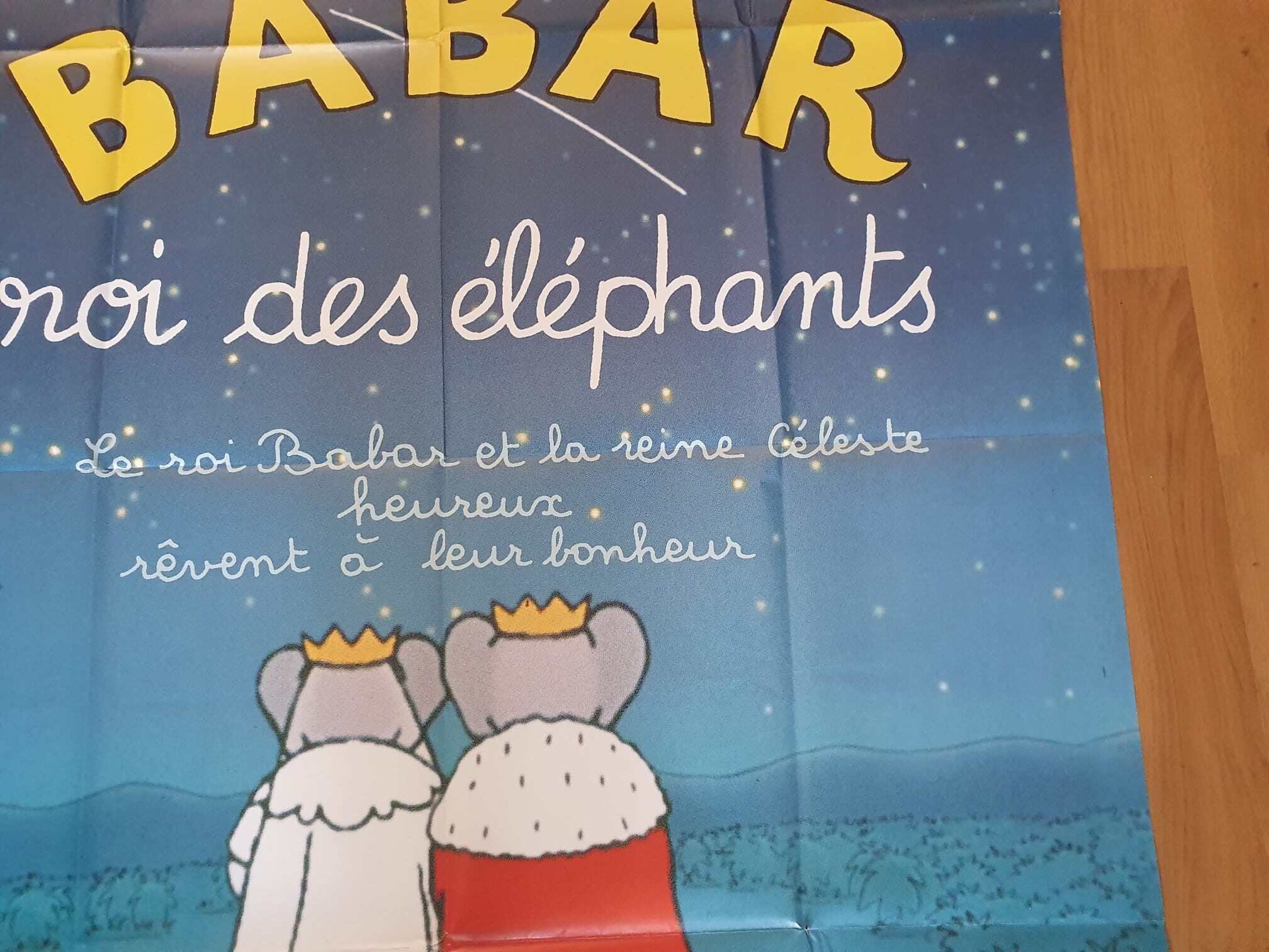 Bien sûr ! Voici la phrase réécrite en utilisant les informations de votre produit : L’« Affiche de cinéma Babar, roi des éléphants – 120 × 162 cm » représente deux éléphants de dessin animé vêtus de robes royales, assis côte à côte sous un ciel étoilé, avec un texte en français au-dessus décrivant le roi Babar et la reine Céleste rêvant de bonheur.