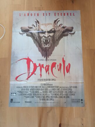Une affiche de cinéma française intitulée « Affiche de cinéma Dracula : L’amour est éternel – 120 × 160 cm » présente, en haut, un visage démoniaque sculpté dans la pierre et un texte en rouge vif. Le générique, la distribution et les détails de production apparaissent sous le titre du film. L’arrière-plan texturé de l’affiche est représenté posé sur un parquet.