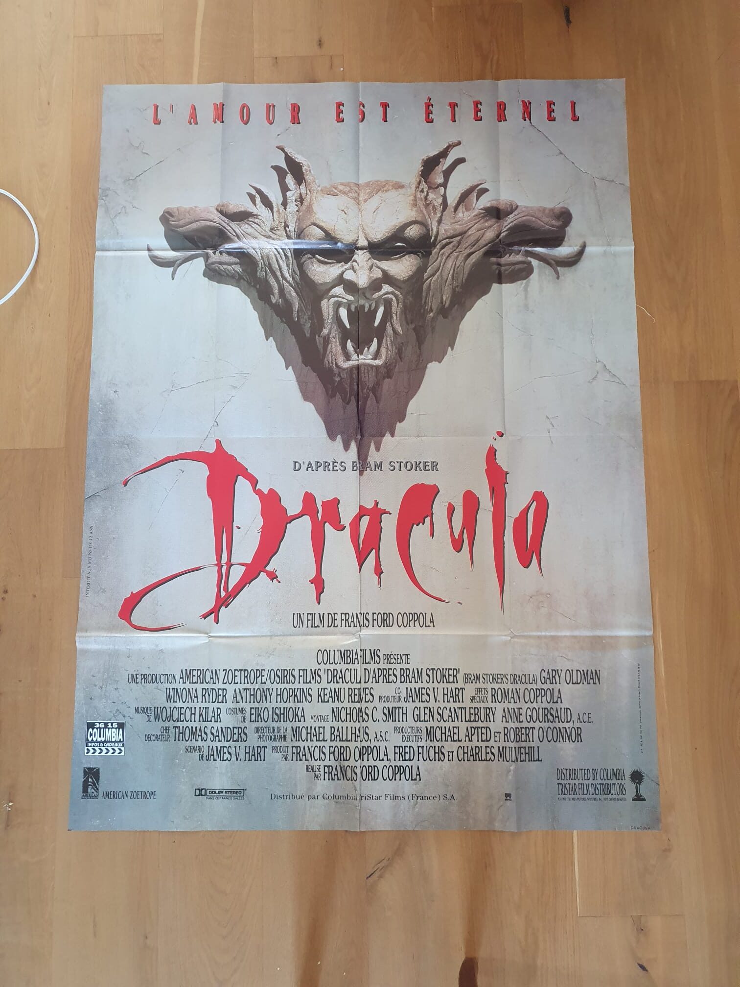 Une affiche de cinéma française intitulée « Affiche de cinéma Dracula : L’amour est éternel – 120 × 160 cm » présente, en haut, un visage démoniaque sculpté dans la pierre et un texte en rouge vif. Le générique, la distribution et les détails de production apparaissent sous le titre du film. L’arrière-plan texturé de l’affiche est représenté posé sur un parquet.
