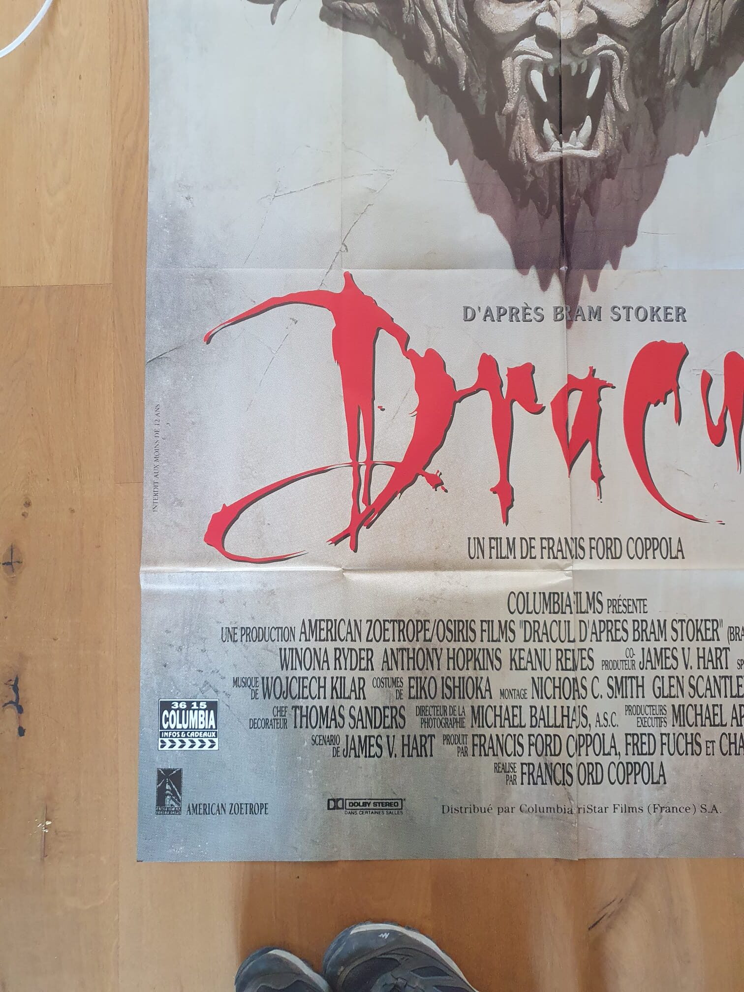 Vue partielle de l’« Affiche de cinéma Dracula : L’amour est éternel – 120 × 160 cm » posée sur un plancher en bois, avec un aperçu des chaussures d’une personne au bas de l’image.