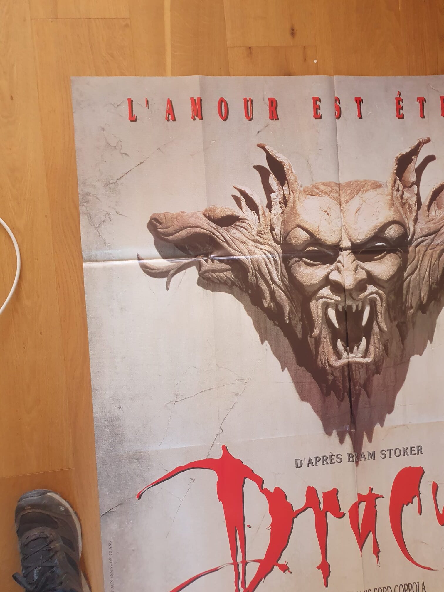 Une affiche de cinéma « Dracula : L'amour est éternel » – 120 × 160 cm – représentant un visage démoniaque en pierre aux crocs acérés, avec un texte en français de couleur rouge et une partie de chaussure visible sur un parquet dans le coin inférieur gauche.
