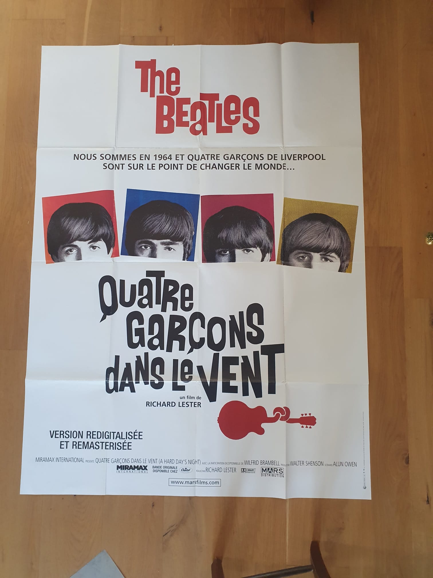 Affiche de cinéma « The Beatles – Quatre garçons dans le vent » (120 × 161 cm), représentant quatre portraits en couleur des membres des Beatles accompagnés d'un texte en français, posée sur un sol en bois.