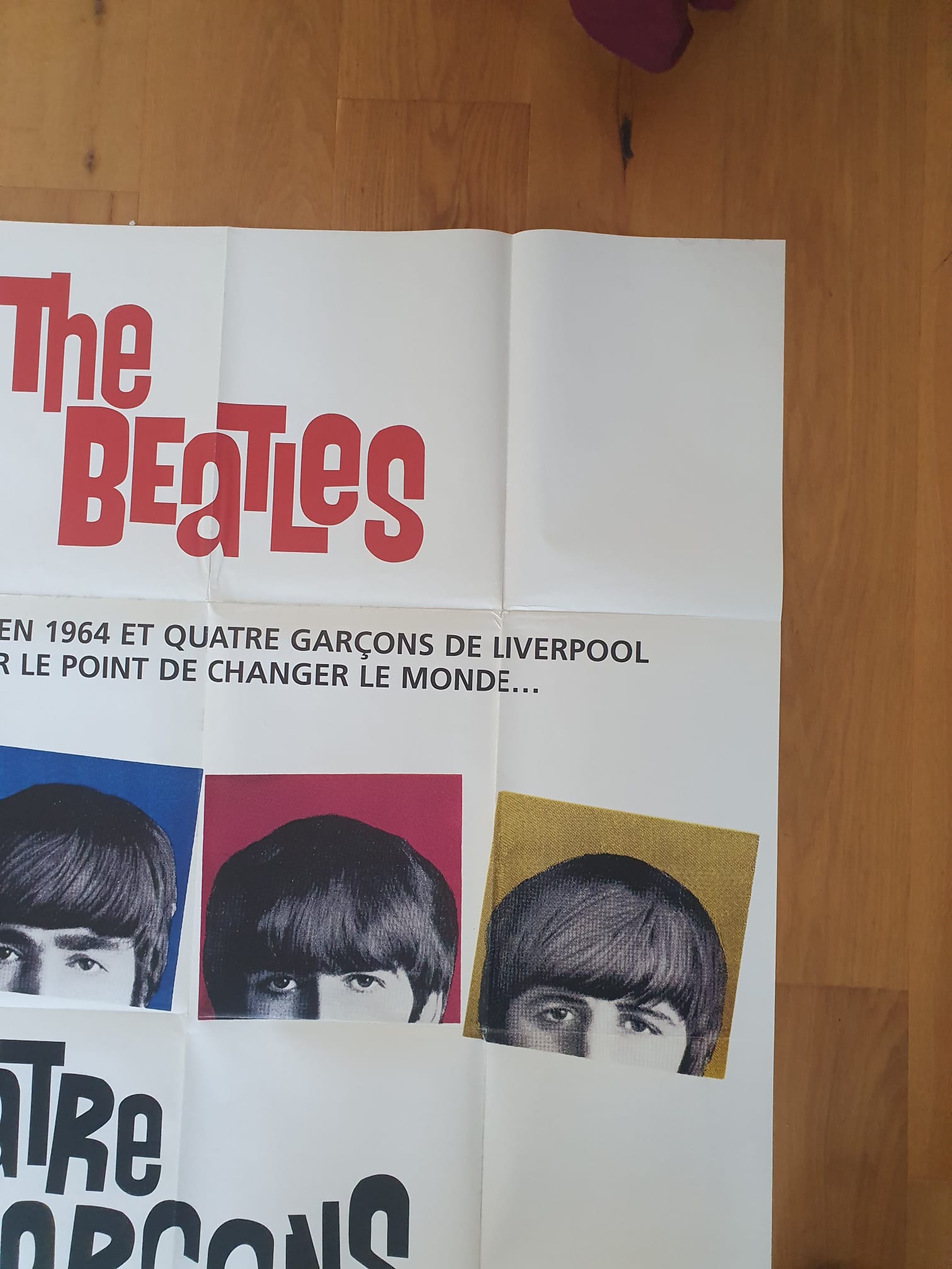 Une affiche dépliée intitulée « Affiche de cinéma The Beatles – Quatre garçons dans le vent – 120 × 161 cm » posée sur un parquet, sur laquelle se détachent des lettrages rouges et noirs audacieux ainsi que des portraits pop art éclatants des membres du groupe, dans des tons bleus, rouges et jaunes.