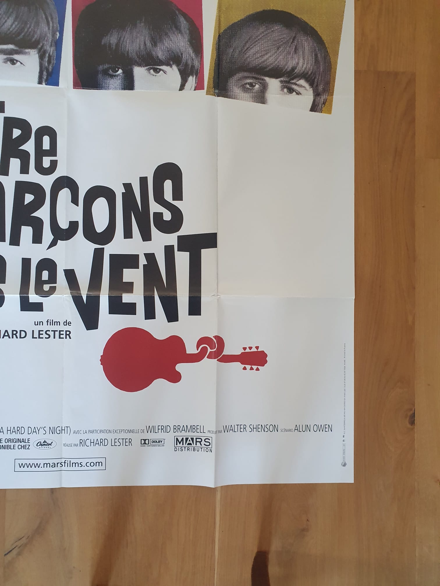 Une affiche de cinéma pliée intitulée *The Beatles – Quatre garçons dans le vent* (120 × 161 cm) repose sur un parquet ; on y voit des visages partiels avec une frange en haut, une inscription en lettres françaises bien en relief, l'illustration d'une guitare rouge au centre, ainsi que le générique et l'adresse d'un site web en bas.