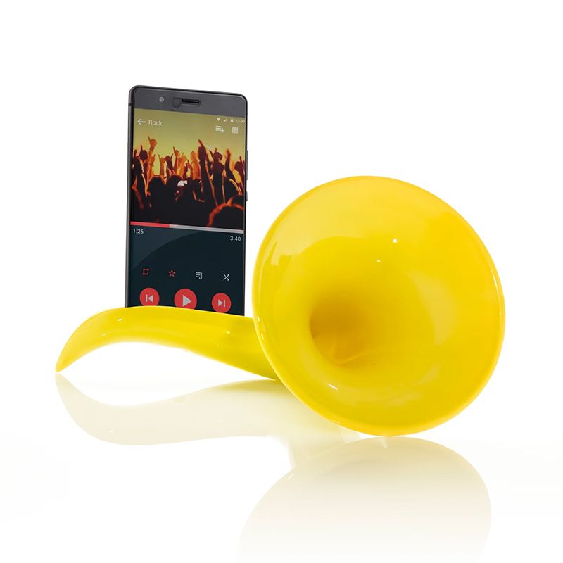 Un amplificateur acoustique « Scaramaz Basic » en céramique jaune, destiné aux smartphones, soutient un appareil diffusant de la musique, dont l'écran affiche une foule de spectateurs lors d'un concert, le tout sur fond blanc.