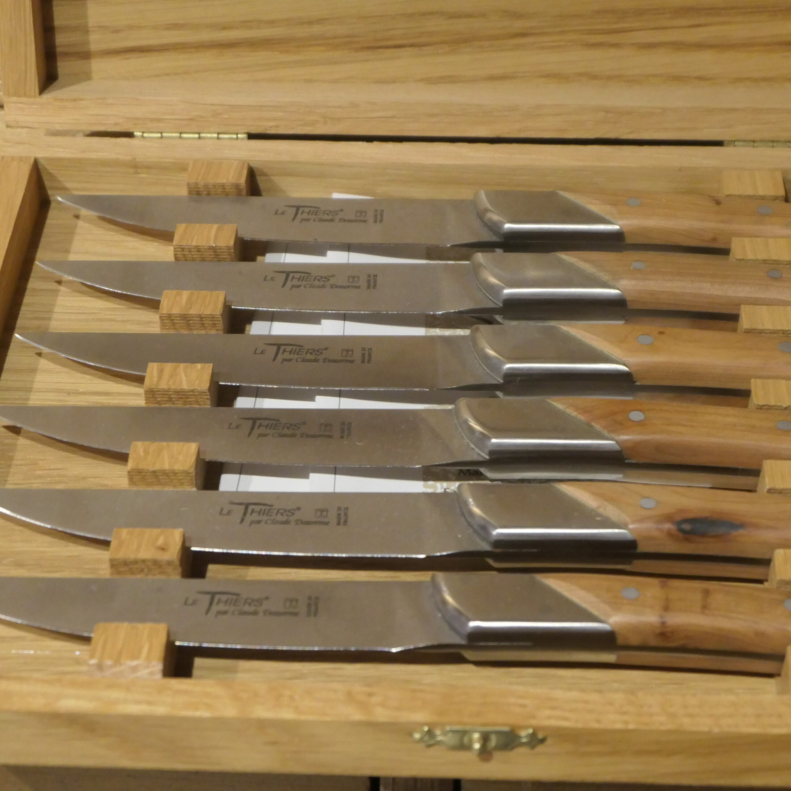Six couteaux de table « Claude Dozorme Le Thiers » sont soigneusement rangés dans un coffret en bois clair, chacun maintenu en place par des séparateurs en bois. Les lames brillantes reflètent la lumière, et le coffret partiellement ouvert laisse entrevoir sa charnière.