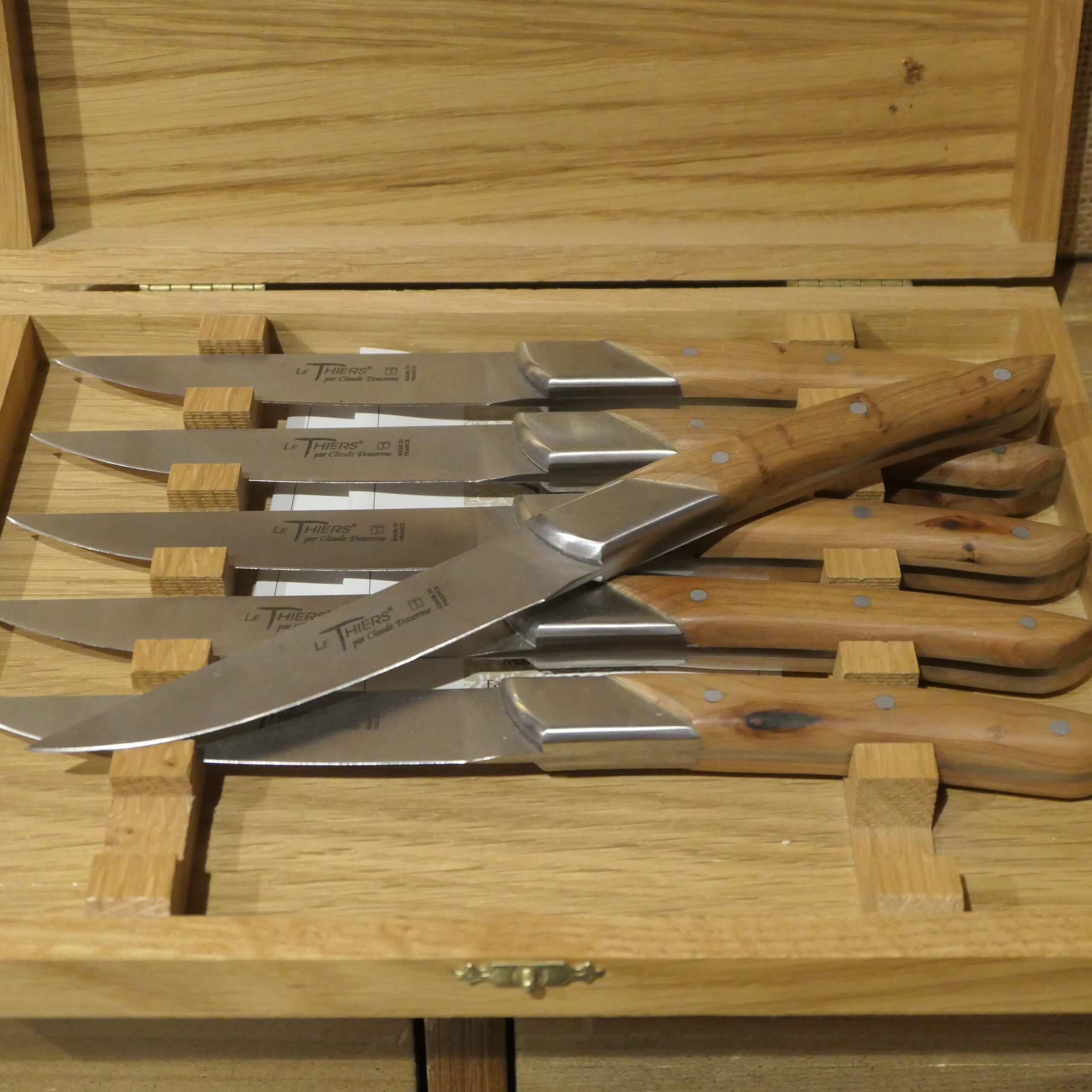Un ensemble de six couteaux de table « Le Thiers » de Claude Dozorme, dotés de manches en bois, est soigneusement rangé dans un coffret en bois comportant des emplacements individuels, toutes les lames étant orientées vers la gauche. Le couvercle ouvert du coffret permet d’admirer les couteaux parfaitement alignés à l’intérieur.