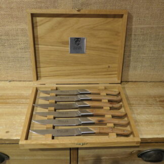 Une boîte en bois contenant six couteaux de table, chacun doté d'un manche en bois et soigneusement rangé dans un emplacement individuel. À l'intérieur du couvercle, une plaque métallique porte le logo Claude Dozorme et l'inscription « Claude Dozorme Le Thiers ».