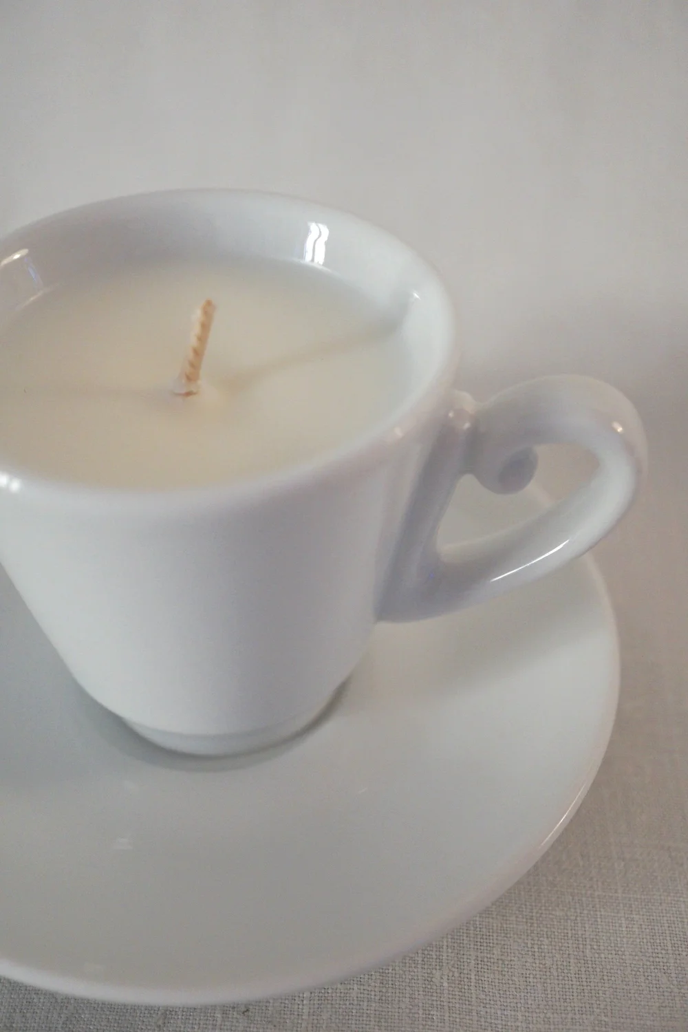 Une « Candela in tazzina - Festina Lente Milano », une bougie en forme de tasse à thé blanche avec une mèche placée au centre, repose sur sa soucoupe assortie, devant un fond clair et neutre.