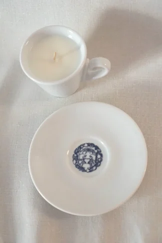La « Candela in tazzina » de Festina Lente Milano, une tasse à thé blanche contenant une bougie blanche, repose sur une soucoupe blanche assortie ornée d’un motif décoratif bleu très travaillés en son centre, le tout disposé sur une surface légèrement texturée.