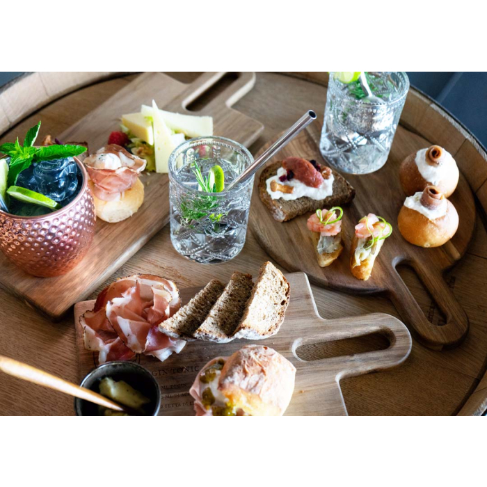 Une table ronde en bois, garnie d'amuse-bouches variés, de tranches de pain, de fromages et de charcuterie, est élégamment disposée sur la « Planche à découper en bois d'acacia FSC® avec recette de cocktail gravée – 50 cm », accompagnée de cocktails agrémentés d'herbes aromatiques et de citron vert, servis sur cette élégante planche en bois d'acacia.