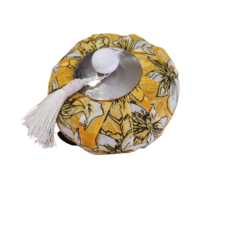 Un récipient rond recouvert de tissu à motifs floraux jaunes, surmonté d'un petit couvercle en métal, d'un bouton blanc et d'un pompon blanc.